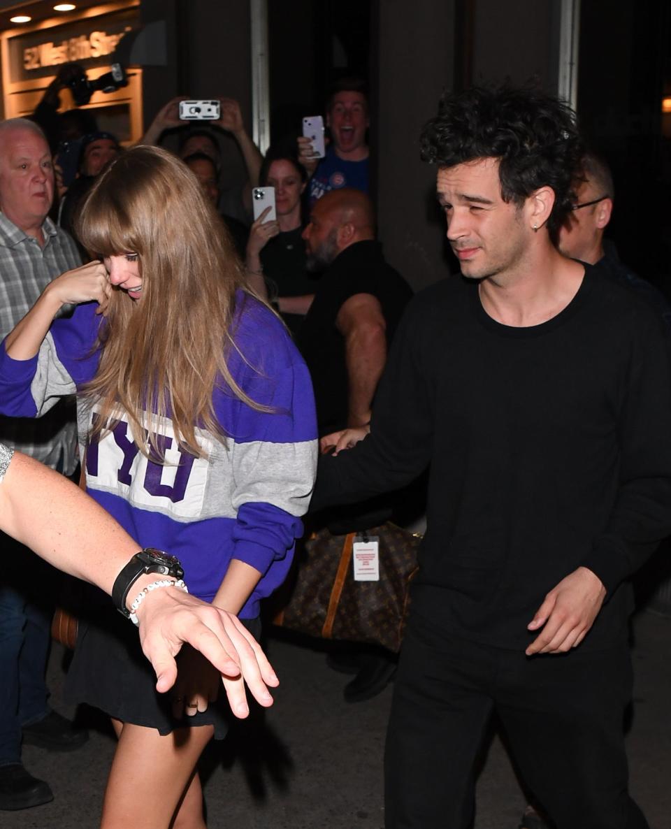 Taylor Swift in a purple sweatshirt with the number "13" escorted by Matty Healy in black through a crowd