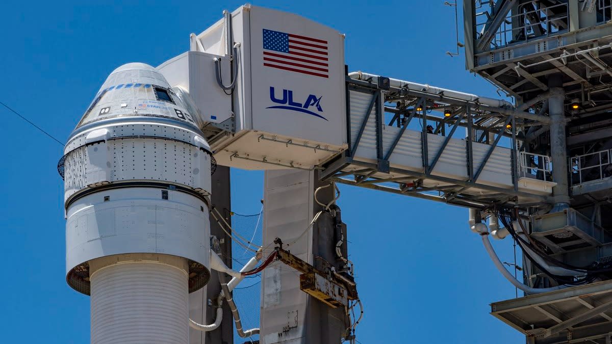  A rocket with a cone-shaped spacecraft on top. it is beside a launch tower. a crew access arm reaches from the tower to the spacecraft with the word "ULA" and an american flag on the side. 