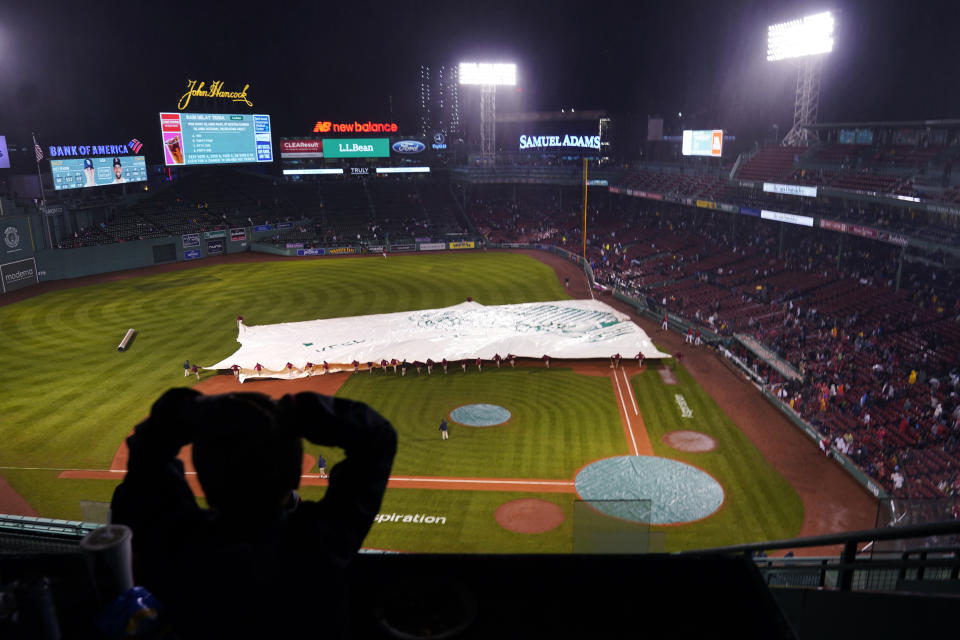 A fan watches as the grounds crew covers the infield as rain falls during the fifth inning of a baseball game between the Boston Red Sox and Tampa Bay Rays, Tuesday, Oct. 4, 2022, at Fenway Park, in Boston. (AP Photo/Charles Krupa)