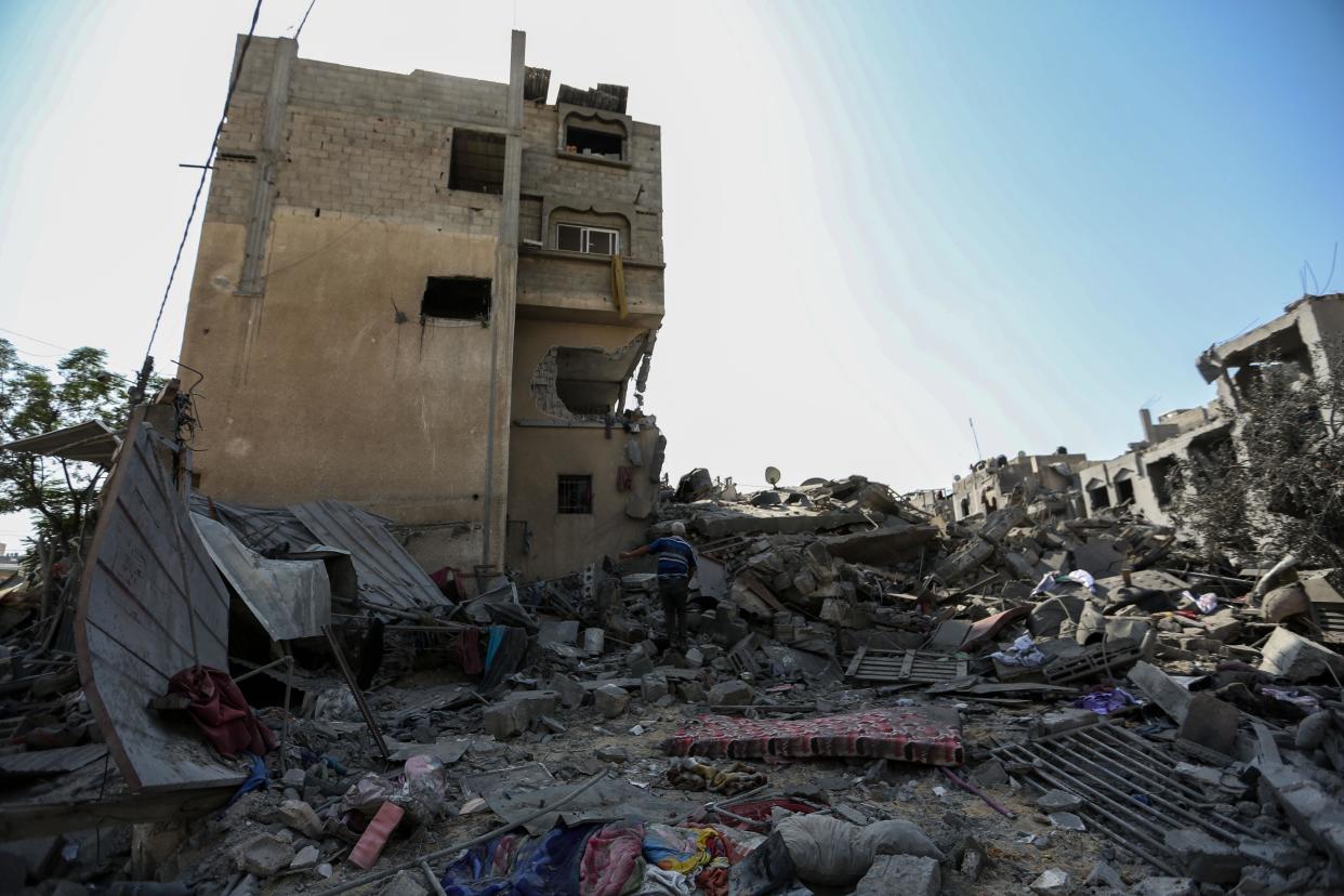 The rubble of grey cement buildings that appear to have been apartments. In the distance, an older man walks through the fallen buildings, putting his hand on one that is still standing.