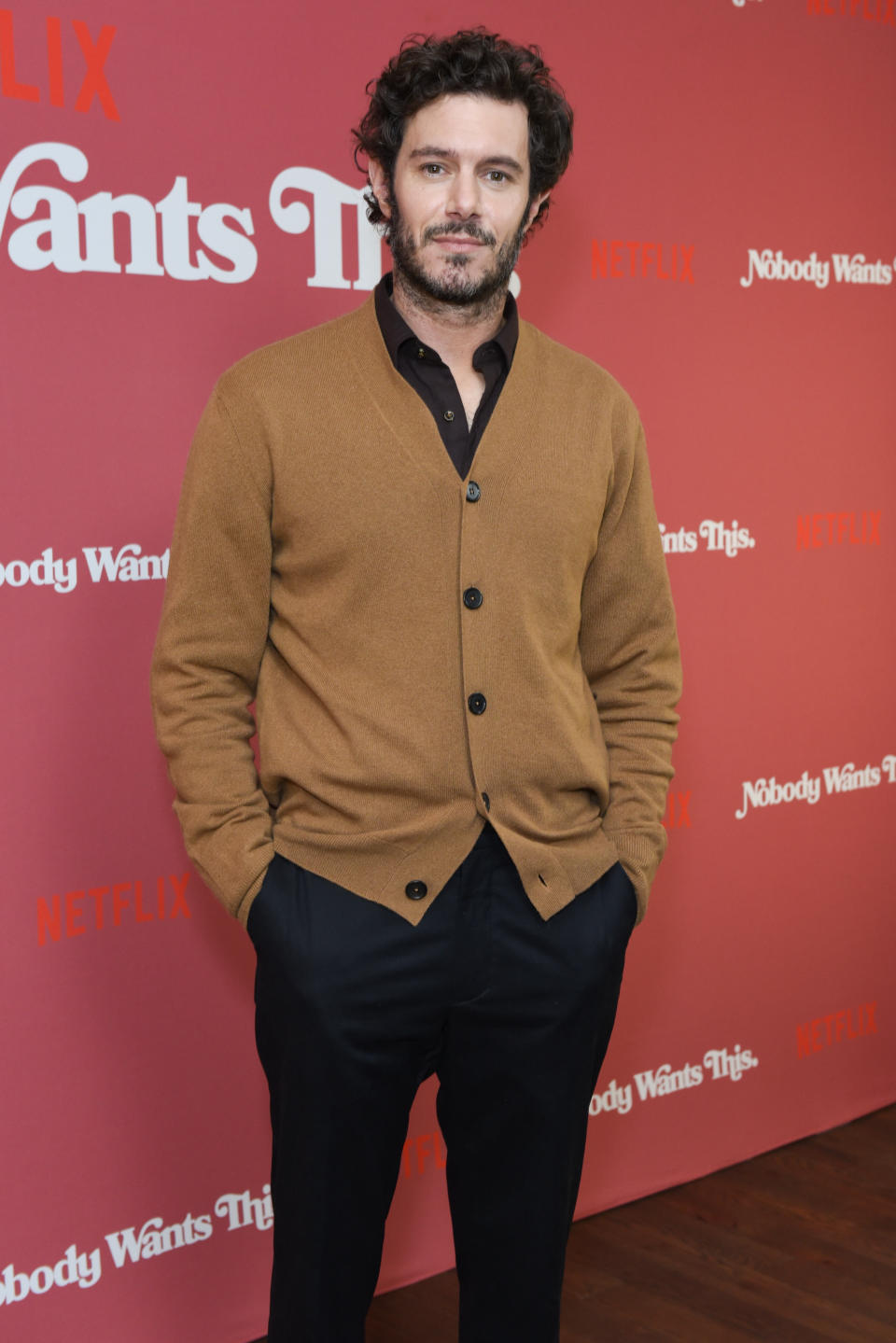 A person stands on a red carpet wearing a brown cardigan and black pants at a Netflix event backdrop with the words "Nobody Wants This."