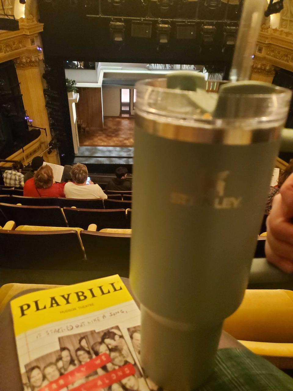 A teal Stanley cup sits atop a Playbill program for Merrily We Roll Along, in a Broadway theater.