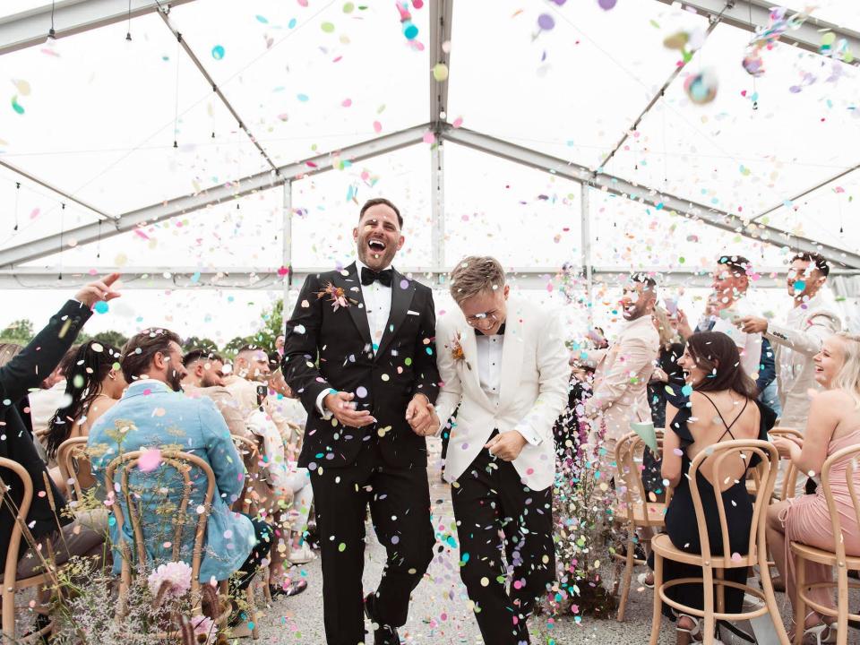 Two grooms laugh as their wedding guests throw confetti on them as they leave their wedding.