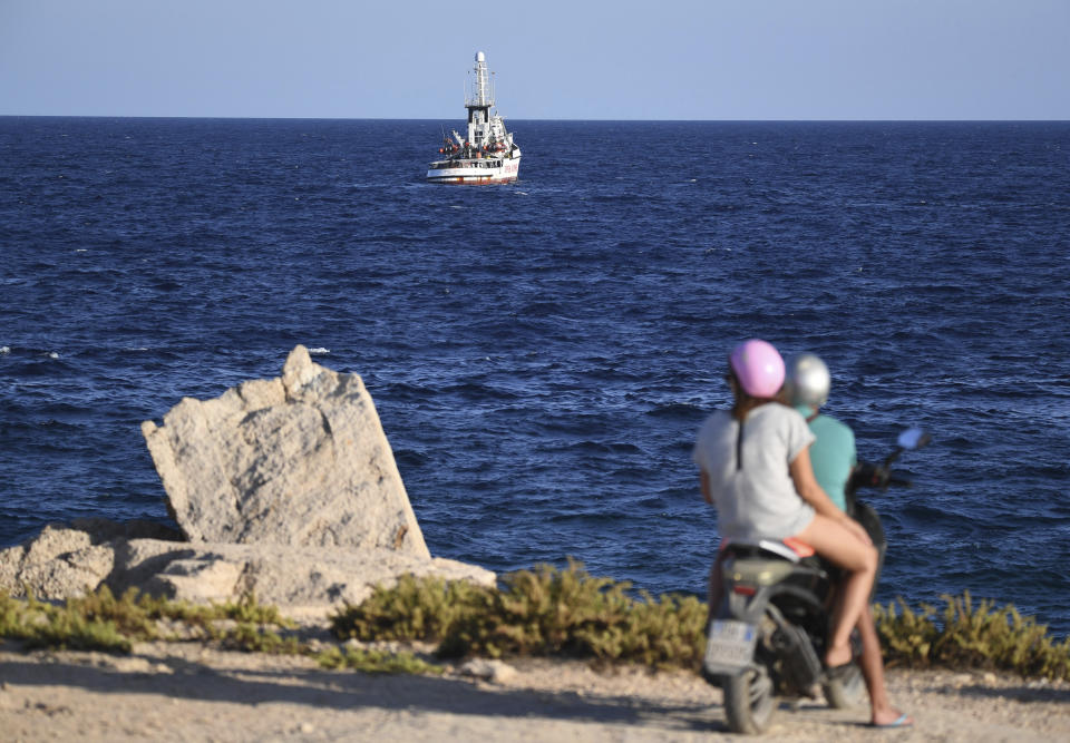 Two people on a scooter stop to look out at the Open Arms vessel, with 107 migrants on board, anchored off the Sicilian vacation and fishing island of Lampedusa, southern Italy, Monday, Aug. 19, 2019. Open Arms on Monday suggested chartering a plane to fly to Spain the migrants blocked off the coast of Italy aboard its boat since early August, to end a stalemate with the Italian Interior minister Matteo Salvini, who won't let private rescue boats into his nation's ports. (AP Photo/Salvatore Cavalli)