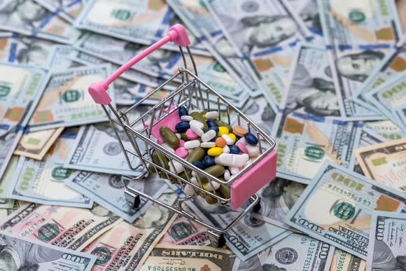 Pink toy shopping cart full of medicine on a pile of cash.