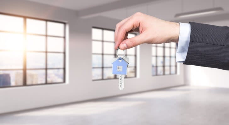 hand of person in a suit dangling keys with a house symbol on the ring. Windows overlooking city skyline in background.