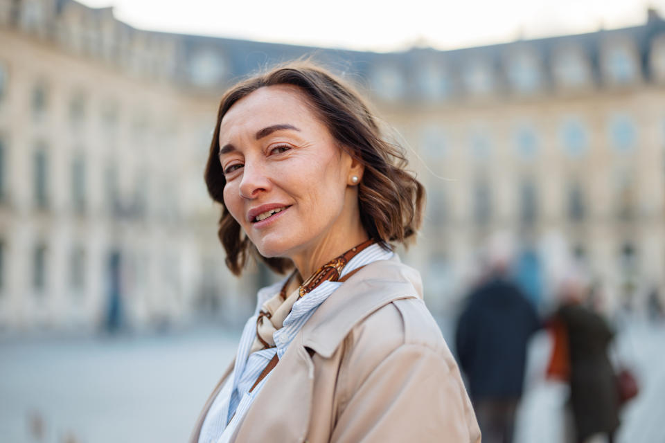 A woman in a city square with historic buildings in the background, wearing a light coat and a scarf