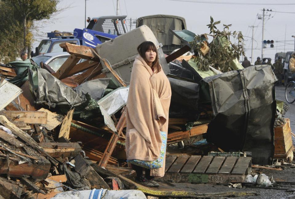 In this March 13, 2011 file photo, a woman named Yuko Sugimoto, wrapped up in a blanket stands in the middle of rubble, looking at the city submerged under water in Ishinomaki in Miyagi Prefecture (state), two days after the catastrophic earthquake-triggered tsunami hit the northeastern Japan. (AP Photo/The Yomiuri Shimbun, Tadashi Okubo, File)