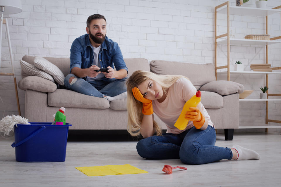 Chauvinism and sexism at home concept. Depressed young lady housewife in rubber gloves holds bottle of cleaning supply, sits on floor, excited husband plays game with gamepad on couch in living room
