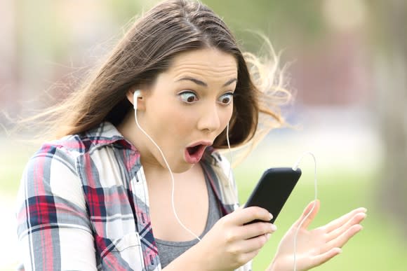 Young woman with headphones staring in wide-eye awe at her smartphone.