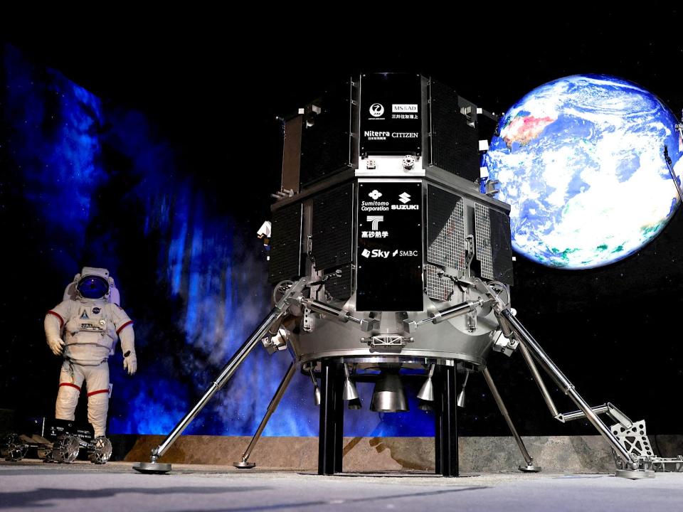 replica of HAKUTO-R lunar lander tall black robot with legs on a stage in front of human spacesuit and a picture of earth