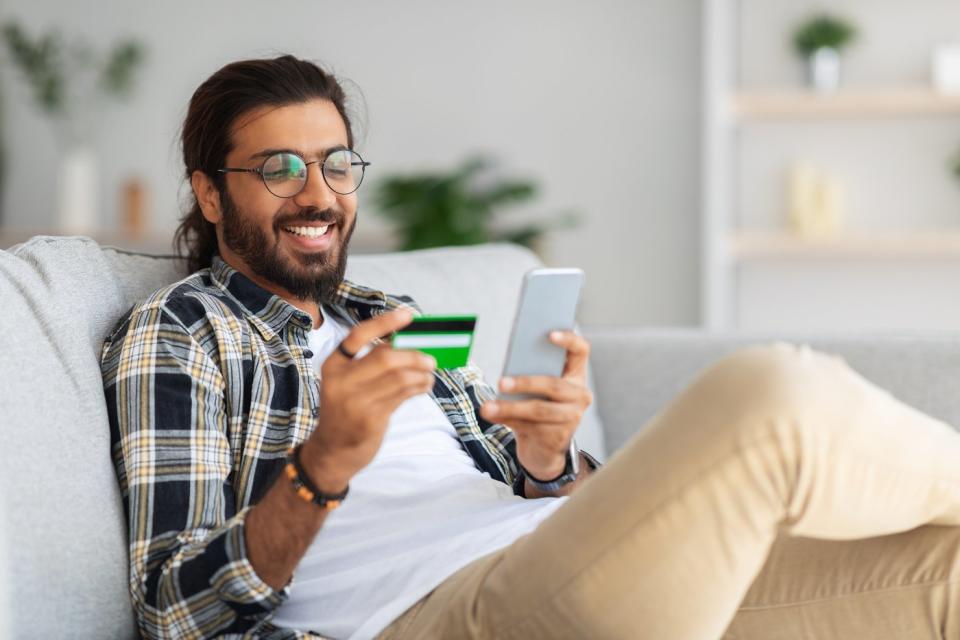 A person in a living room holds up a credit card and looks at a phone.