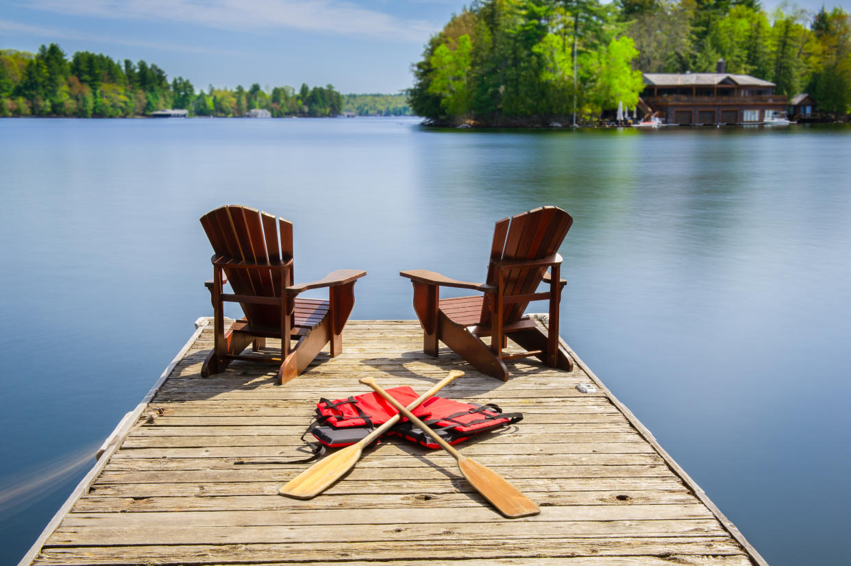 Two Muskoka chairs on a wooden dock facing a lake. Paddles and life jackets are visible on the dock. Across the calm water there is a brown cottage.