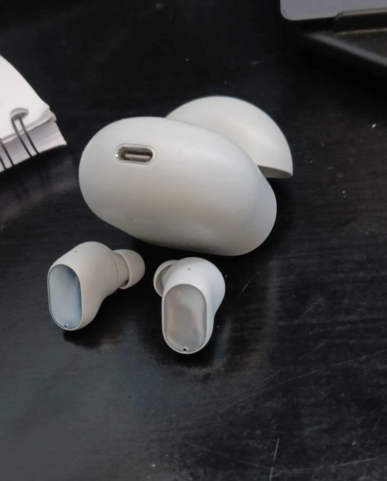 Close-up of wireless earbuds and their charging case on a dark surface next to a notebook, looking like someone in a sexual position