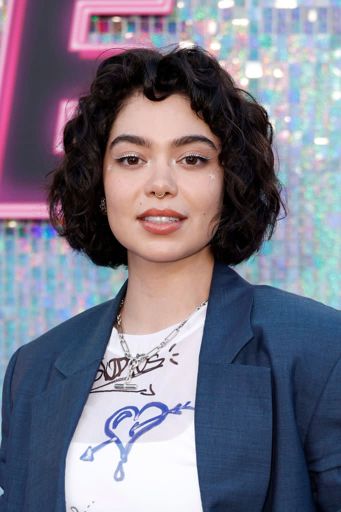 Auli'i Cravalho at a public event, wearing a graphic white shirt, a dark blazer, and layered necklaces. She has shoulder-length curly hair and minimal makeup