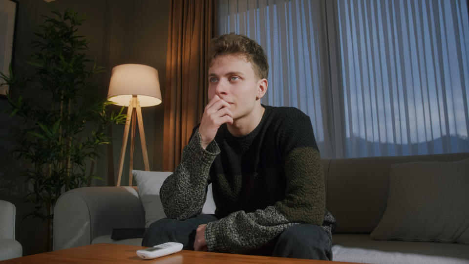 Handsome young man sits on the couch at evening at home, watching a movie on TV