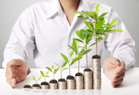 A person standing behind rising stacks of coins with growing plants on each one.