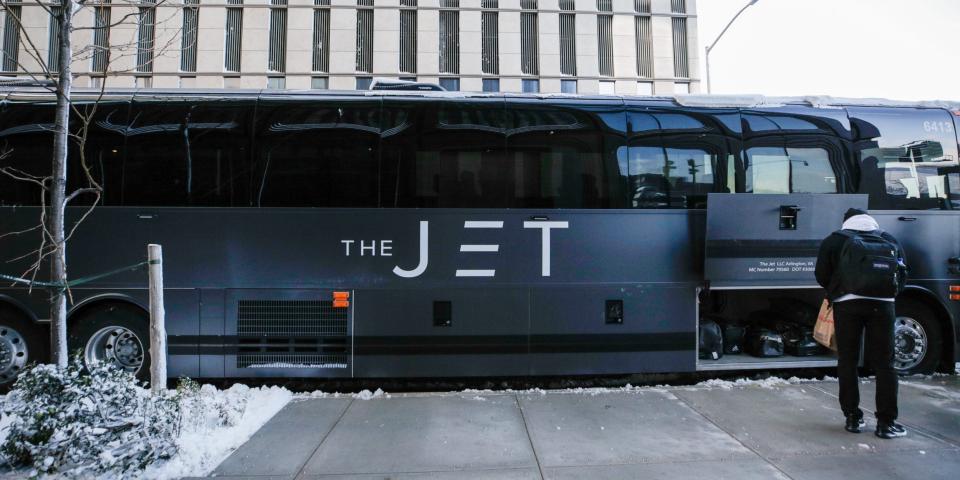 A matte black bus that reads "The Jet" on the side. Passengers with bags are boarding the bus or putting their bags away into the lower storage compartment.