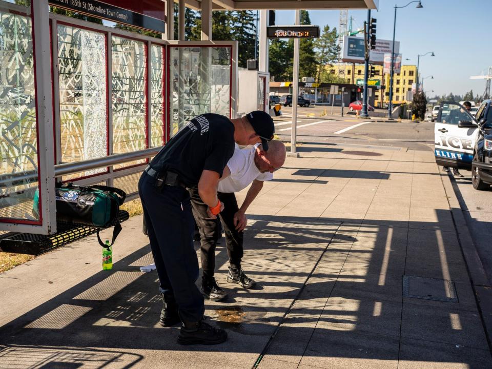 emt in uniform talks to man bending over with hands on knees at a bus stop on the sidewalk