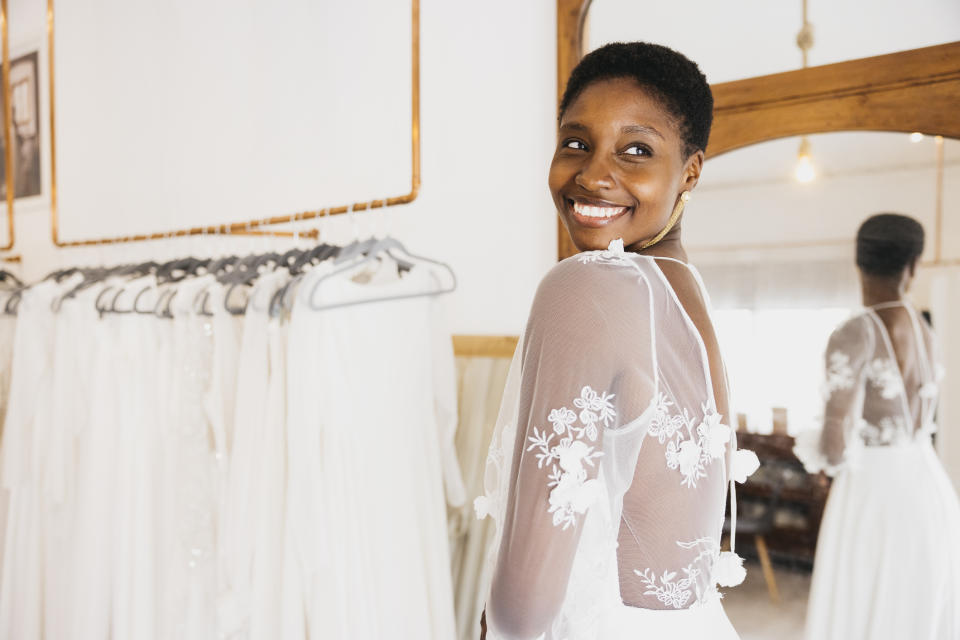 A bride smiles joyfully while wearing a wedding dress with floral lace details on the back. She stands in front of a mirror in a room with other dresses hanging