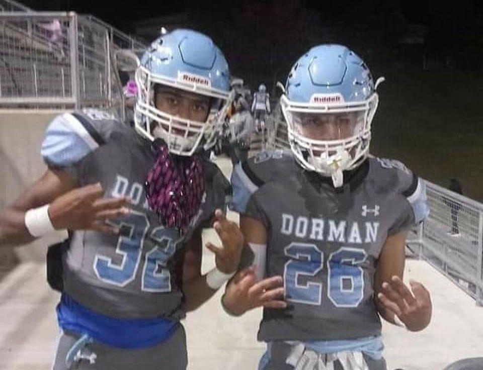 Keegan Johnson (wearing a shirt saying Dorman 26) and Nate Worthy (32), suited up with their football helmets and both touching their index fingers to their thumbs, are pictured at night.