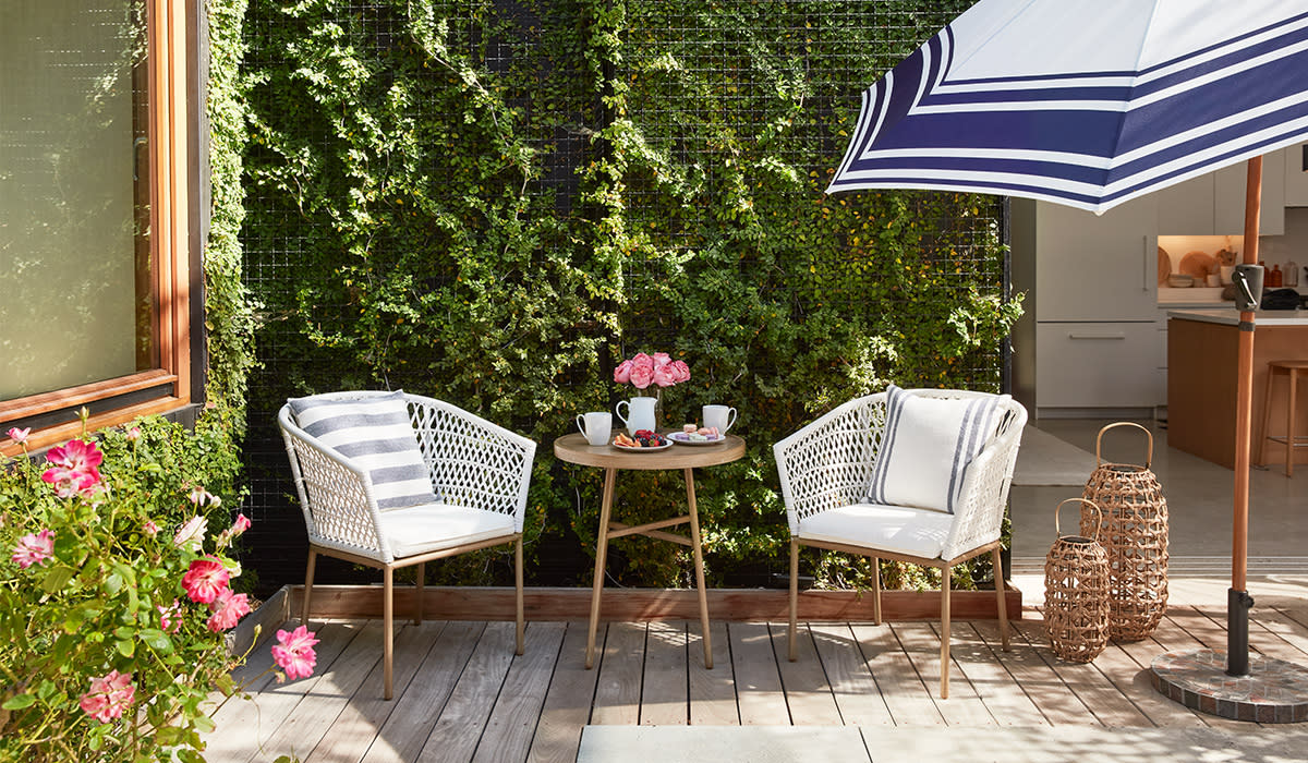 Pair of chairs beneath striped umbrella with coffee and fruit on table between them. 