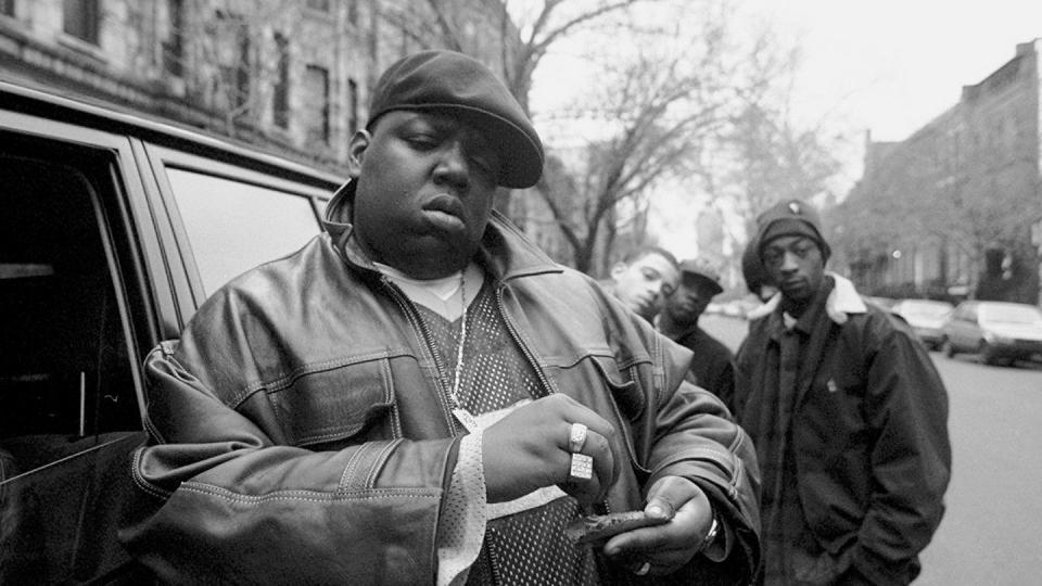 a black and white photo of the notorious big standing next to a car parked on a city street, rolling a cigar, with several people in the background looking at the camera