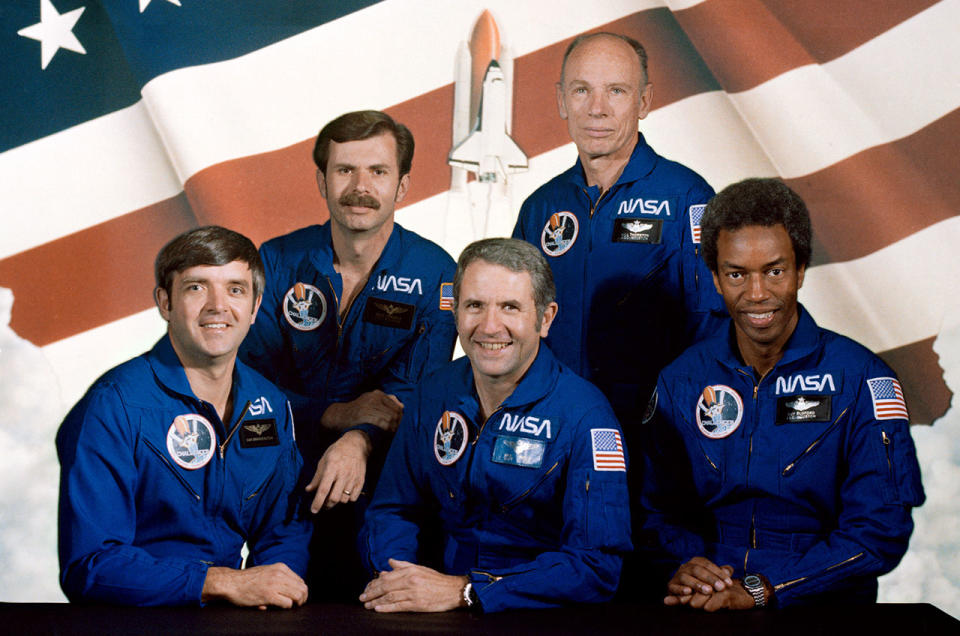 five men in blue flying suits stand in front of an American flag