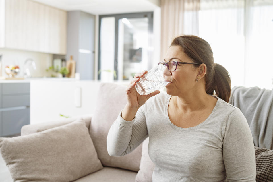 A woman with glasses and a ponytail drinks a glass of water while sitting on a couch in a modern kitchen and living room space