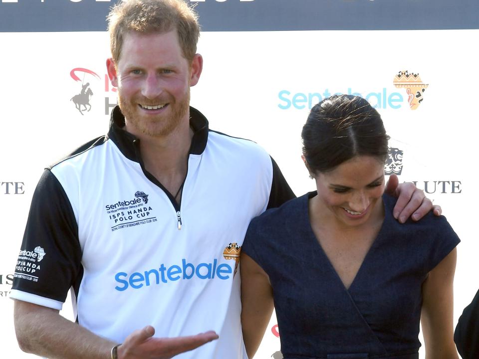 Harry in a polo shirt smiling and pointing to Meghan in a navy dress whose smiling and looking down