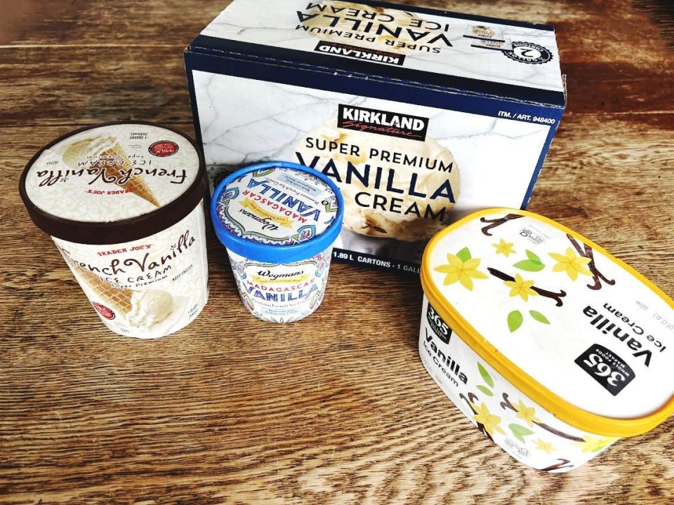 Several cartons of ice cream sit on a wooden table. From left to right, the table holds a white carton of ice cream with Trader Joe's logo and a picture of an ice-cream cone on it, a small pint with a red, green, and blue design and Wegmans logo on the front, a large box with blue, gray, and Kirkland Signature on it, and a white and yellow carton of ice cream with 365 logo on it