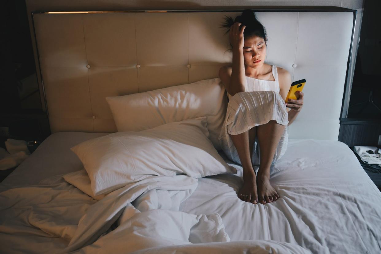 Portrait of depressed woman sitting alone on the bed in the bedroom and looking to mobile phone in her hand.