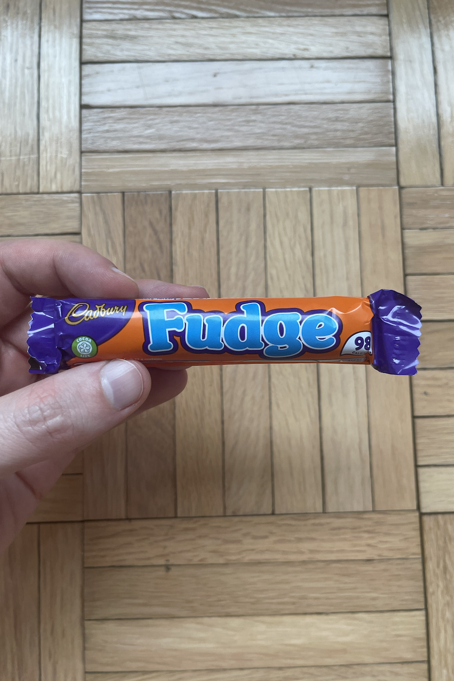 Hand holding a Cadbury Fudge chocolate bar against a wooden floor background