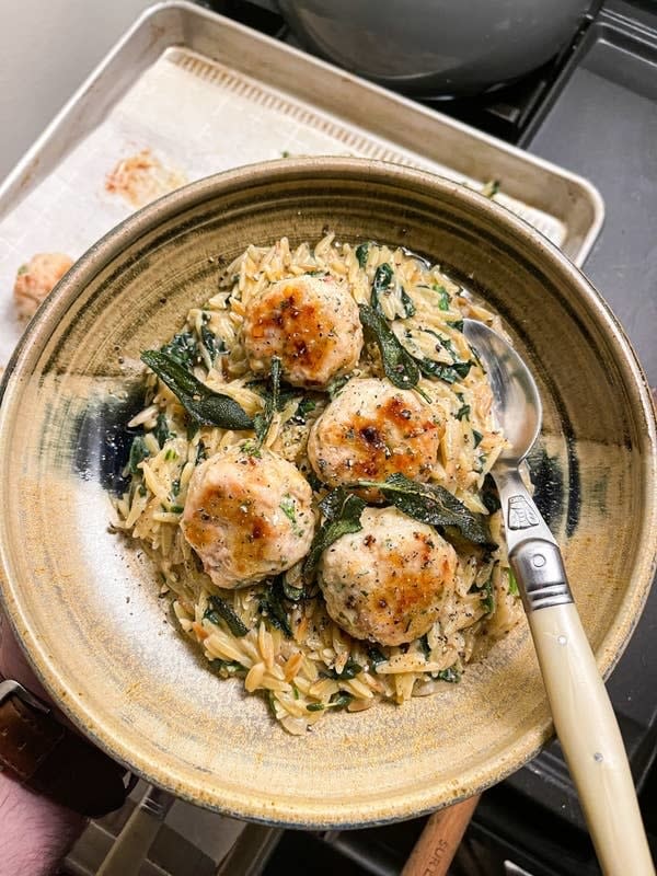 A bowl of orzo pasta with meatballs and spinach, fork on the side; a person's hand visible