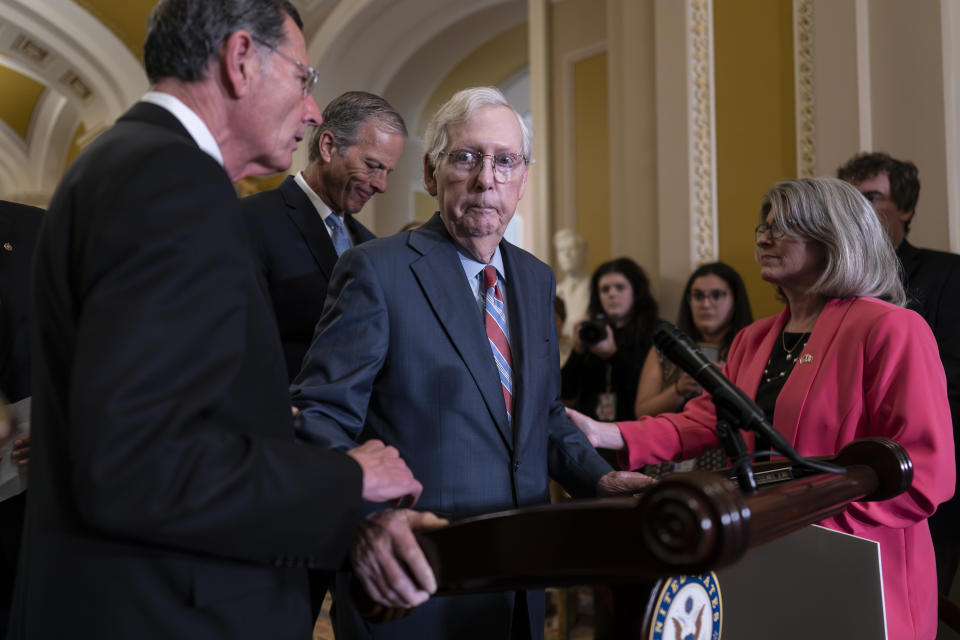 Sen. Mitch McConnell, looking blank, is helped by, on his left, Sen. John Barrasso, Sen. John Thune and Sen. Joni Ernst.