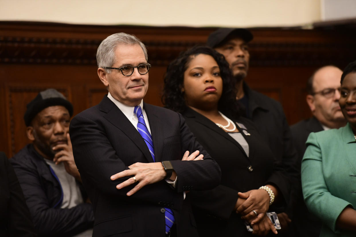 Larry Krasner, in a wood-paneled room surrounded by other people, crosses his arms and wears a faint smile.