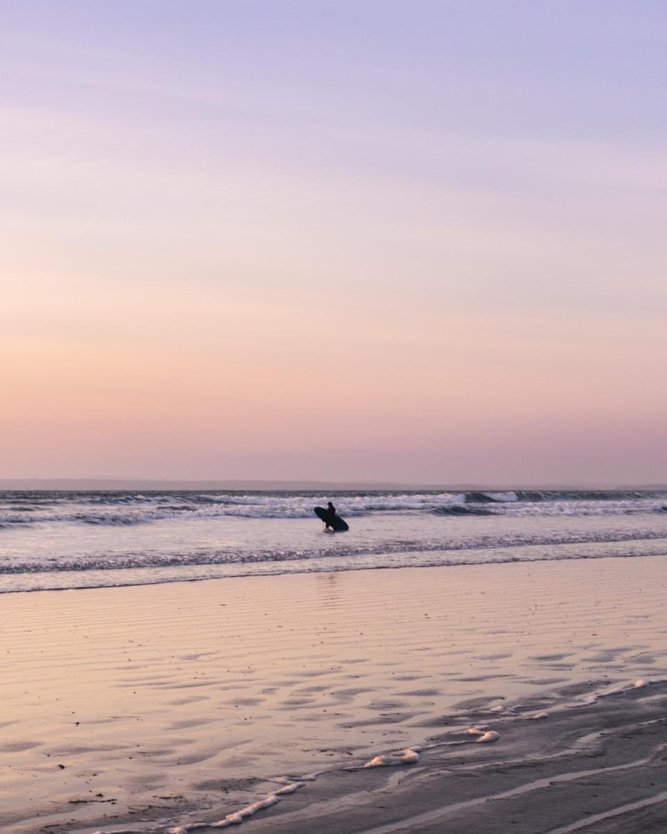 Photo of a distant lone surfer standing in gentle waves on the beach.