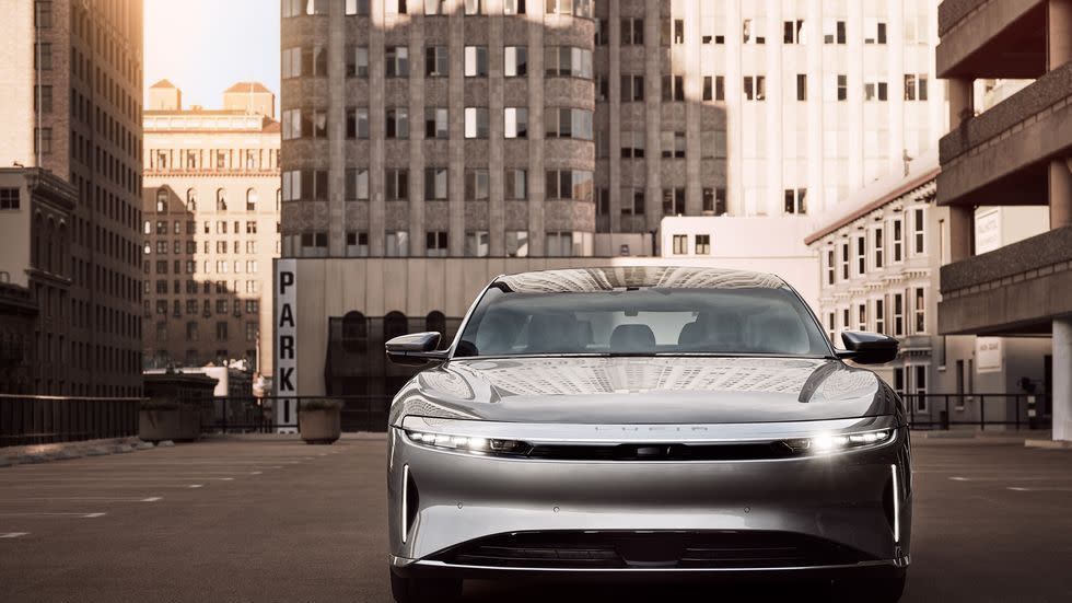 a silver 2023 lucid air touring sedan parked face front in a city yard with tall buildings in the background