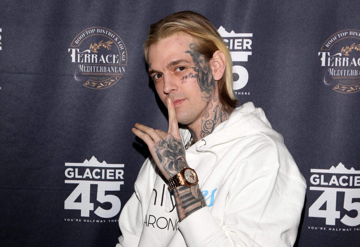 Aaron Carter, the side of his face, neck, hand and wrist covered in tattoos, puts his hand to his lips as he poses for a red carpet photo.