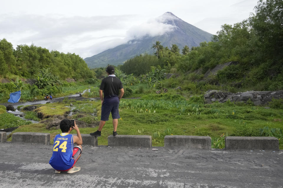 A boy takes a photo of his friend who poses in front of the Mayon volcano in Bonga, Albay province, northeastern Philippines., Saturday, June 10, 2023. Monsoon rains that could be unleashed by an offshore typhoon were complicating worries of villagers threatened by a restive Philippine volcano that has forced thousands of people to flee from their homes.(AP Photo/Aaron Favila)