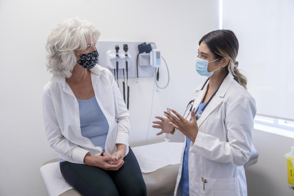 An older woman in a casual outfit and mask sits on a medical examination table while a healthcare provider in a lab coat and mask speaks to her