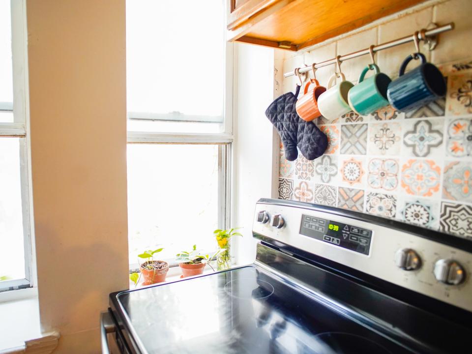 A view of the author's kitchen in her Brooklyn apartment shows an electric stove on the left with mugs hanging on a rack above it and windows behind them.