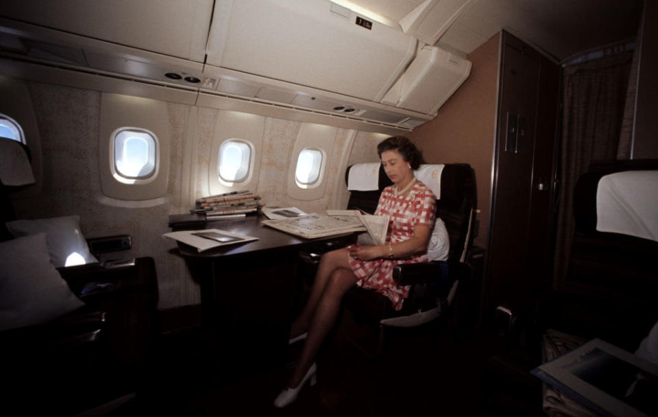 A person in a checkered dress reads a newspaper while seated in an airplane cabin, with documents resting on a table beside them