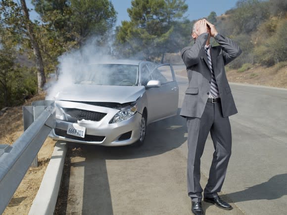 A man in a suit puts his hands on his head while standing in front of a car that crashed into a guardrail.