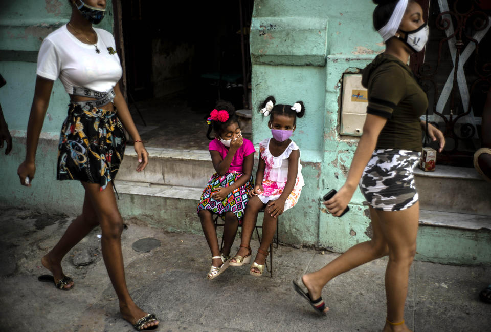 Wearing a mask as a precaution against the spread of the new coronavirus Angelica Victoria, center left, and Thalia Oneida, wait for their parents sitting on a chair in Havana, Cuba, Monday, Oct. 12, 2020. Cuba relaxed coronavirus restrictions Monday in hopes of boosting its economy, allowing shops and government offices to reopen and welcoming locals and tourists at airports across the island except in Havana. (AP Photo/Ramon Espinosa)