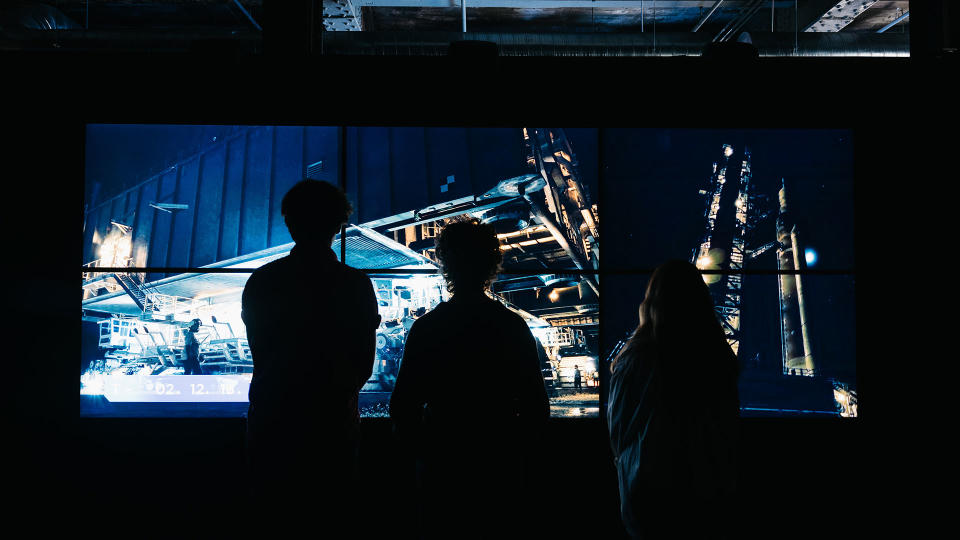 a photo showing three silhouettes in the foreground looking at a projection of a rocket launch tower