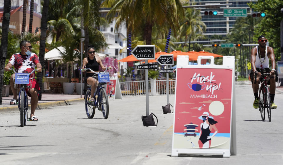 FILE - In this Aug. 11, 2020, file photo, bike riders ride past a "Mask Up Miami Beach," sign, in Miami Beach. As states around the country require visitors from areas with high rates of coronavirus infections to quarantine upon arrival, children taking end-of-summer vacations to hot spots are facing the possibility of being forced to skip the start of in-person learning at their schools. (AP Photo/Wilfredo Lee, File)