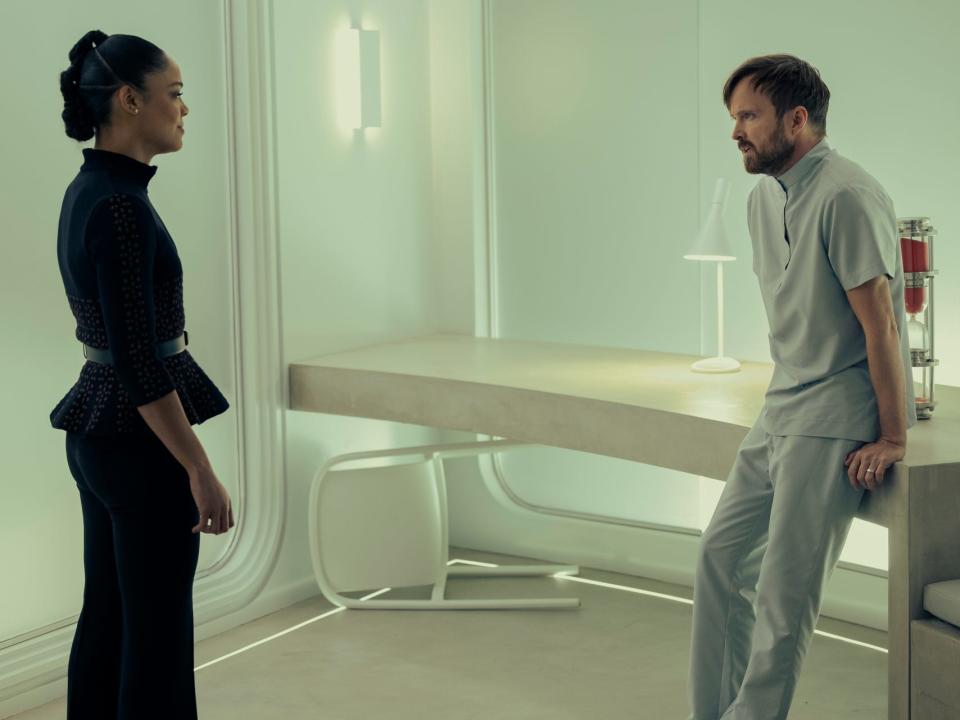 A woman and man stand facing each other in a white room.
