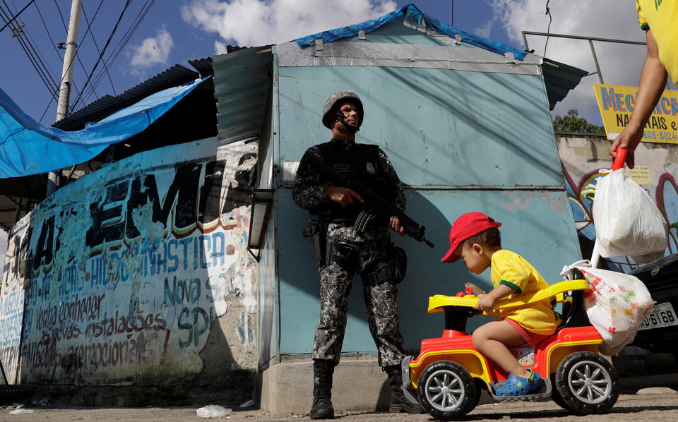 A boy on a cart is pushed near a Brazilian Public-Safety National Force policeman