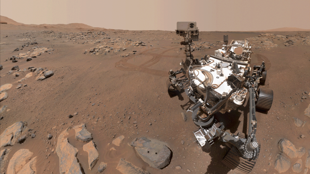  A wheeled robot looks around with its camera-like "head" on a dusty red lake bed strewn with rocks. 